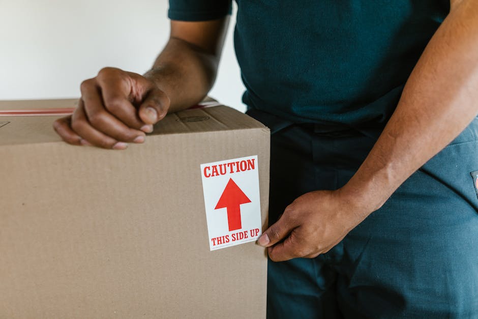 Close-up view of a person in dark clothing handling a cardboard moving box marked with a red and white caution sticker that reads 'Caution This Side Up' with an upward-pointing arrow. The individual i